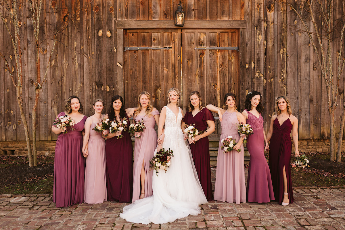 Bride and bridesmaids stand on brick path in front of large wooden barn door