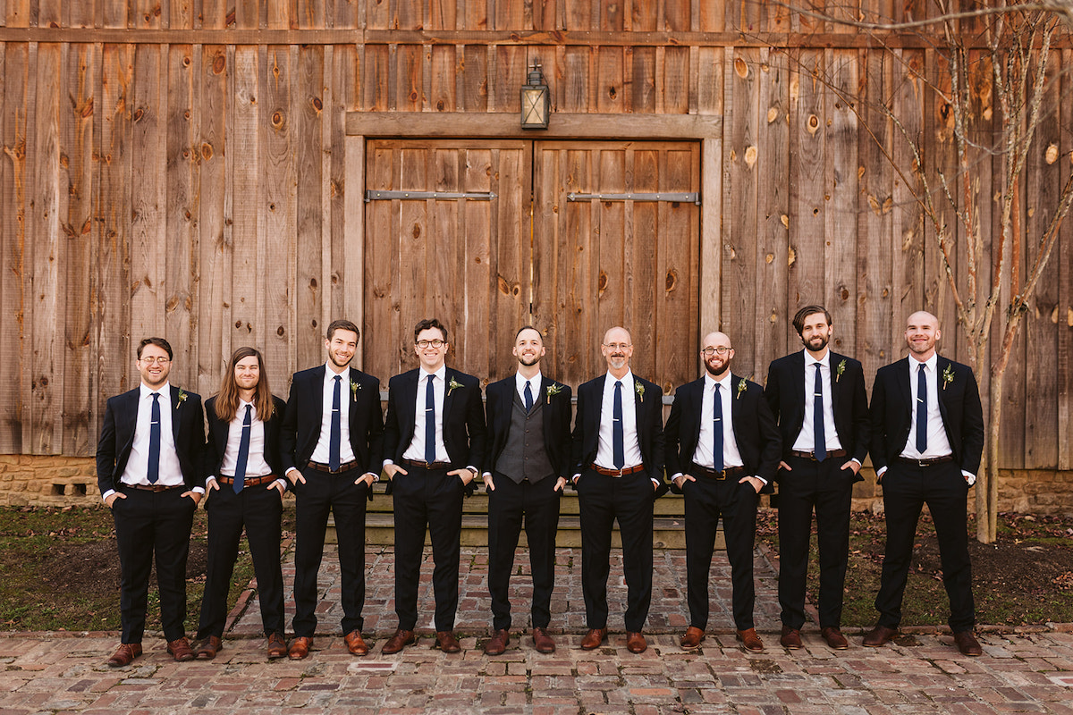 Groom stands with groomsmen in their dark suits in front of tall wooden barn doors