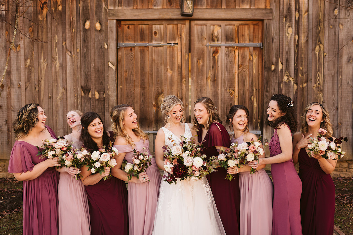 Bride and bridesmaids laugh together in front of large wooden barn door