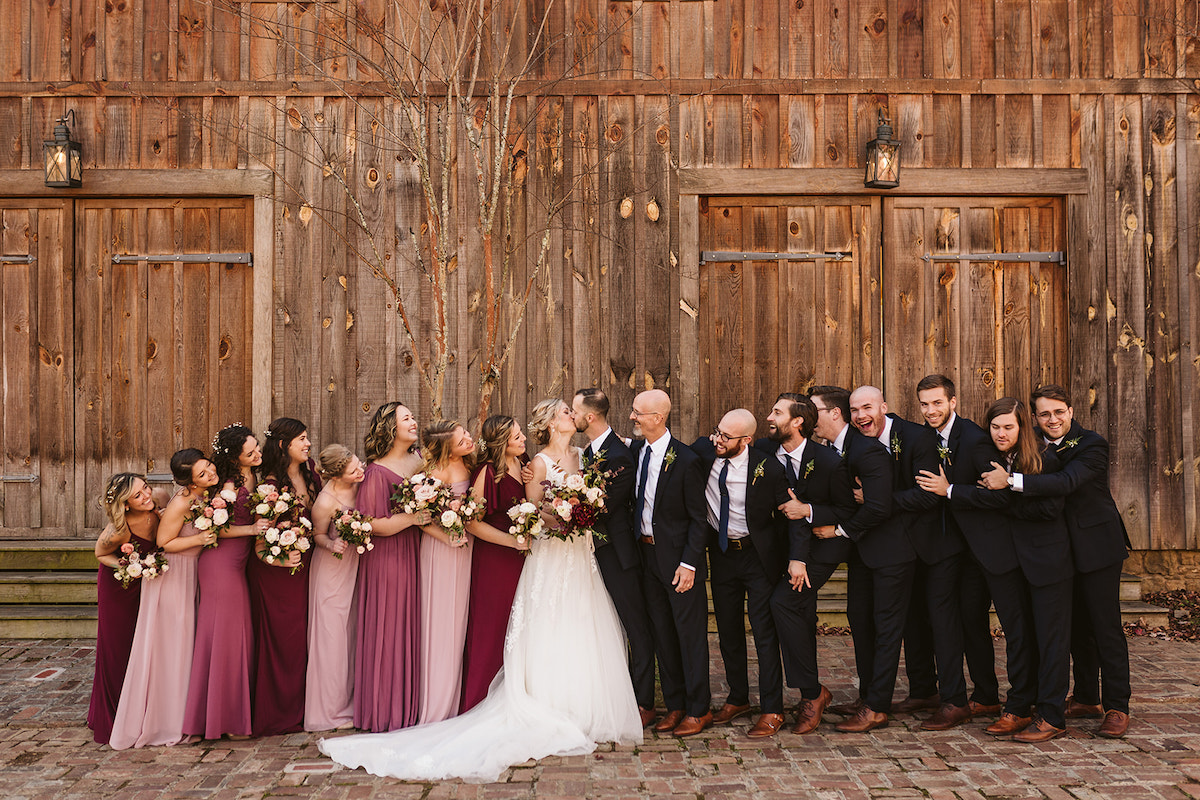 Bride and groom stand with their bridal party on brick path in front of large wooden barn front of tall wooden barn doors