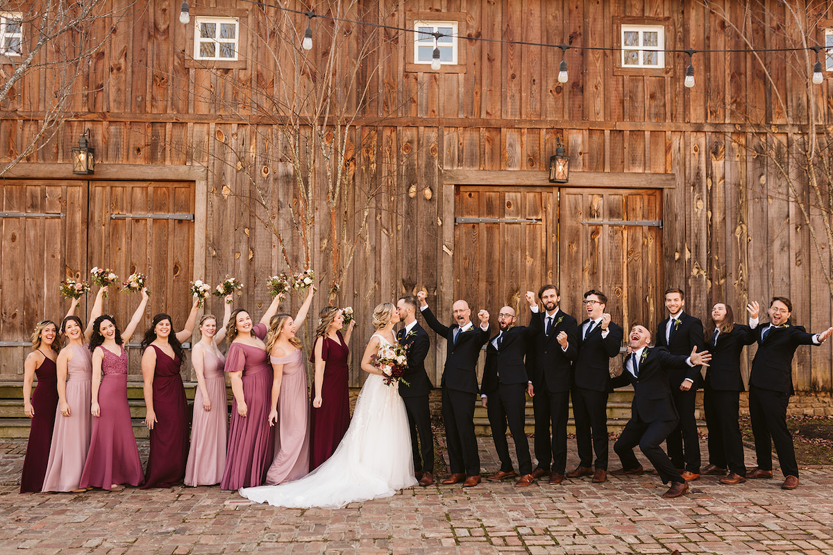 Bride and groom stand with their bridal party on brick path in front of large wooden barn front of tall wooden barn doors