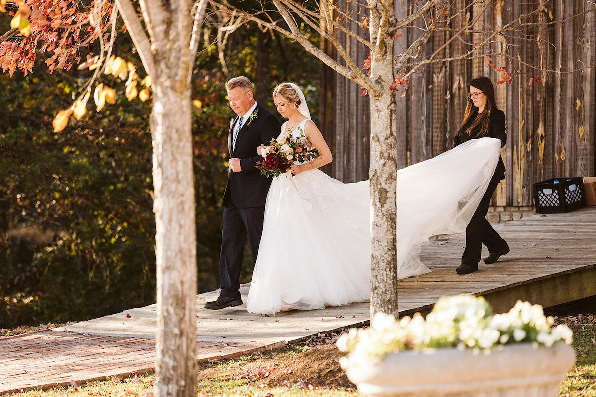 Bride and her father walk down a wide wooden ramp, while Mia + May wedding coordinator fluffs her train behind her