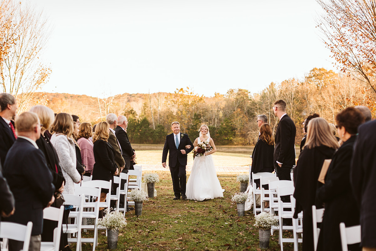 Bride is escorted by her father down the center grass aisle. Guests stand from their white wooden fold-up chairs.