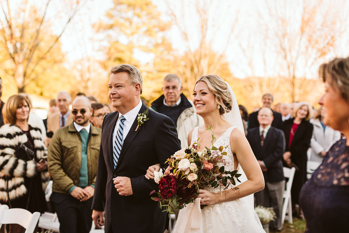 Bride is escorted by her father down the aisle. Guests stand from their white wooden fold-up chairs.