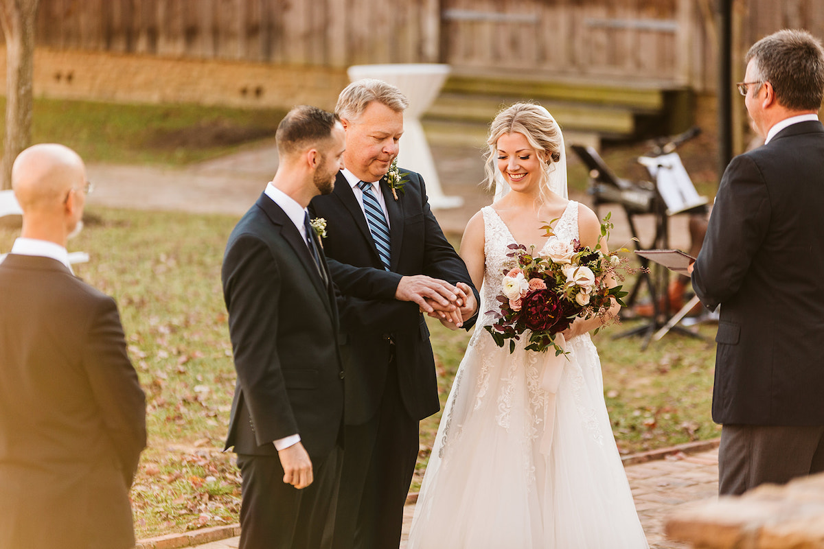 Bride's father joins her hand with groom's hand as she holds her Petaline floral bouquet of deep red and white flowers