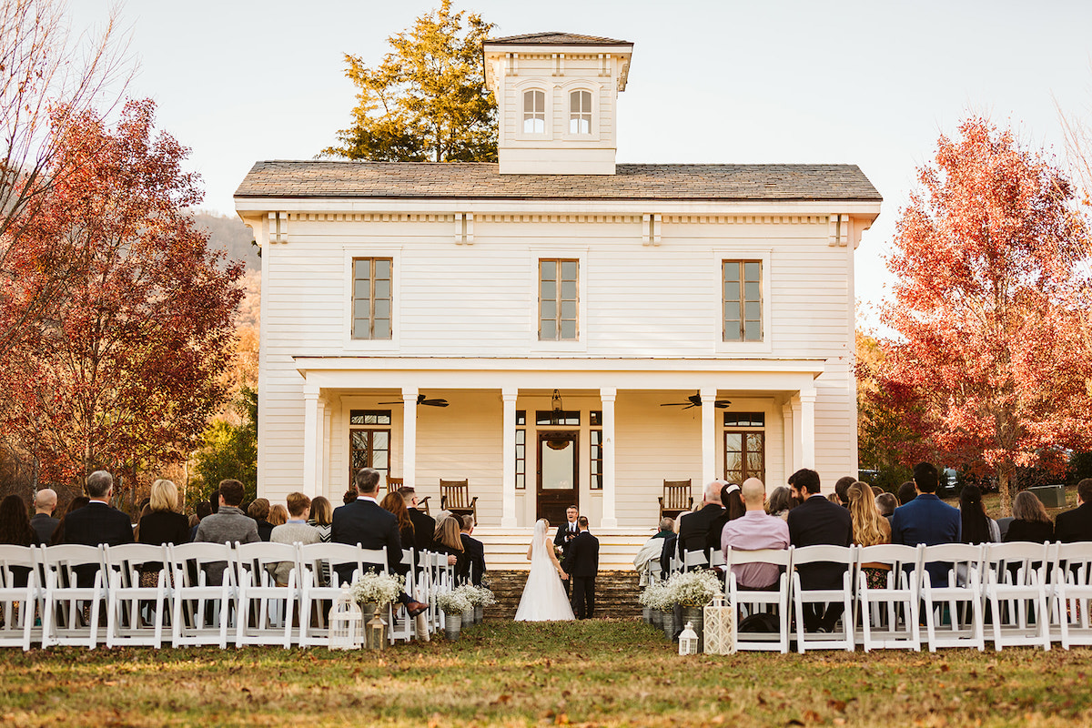 Bride and groom stand at the base of the stone steps in front of Peacock Hall while guests sit in white chairs on the lawn