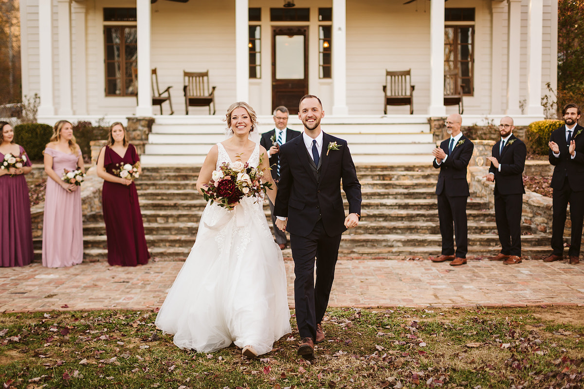 Bride and groom walk hand-in-hand, smiling, while bridal party claps and watches them walk away