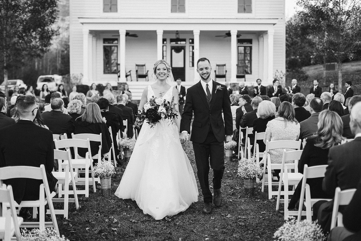 Bride and groom walk down the aisle hand-in-hand, smiling, while guests sit facing Peacock Hall