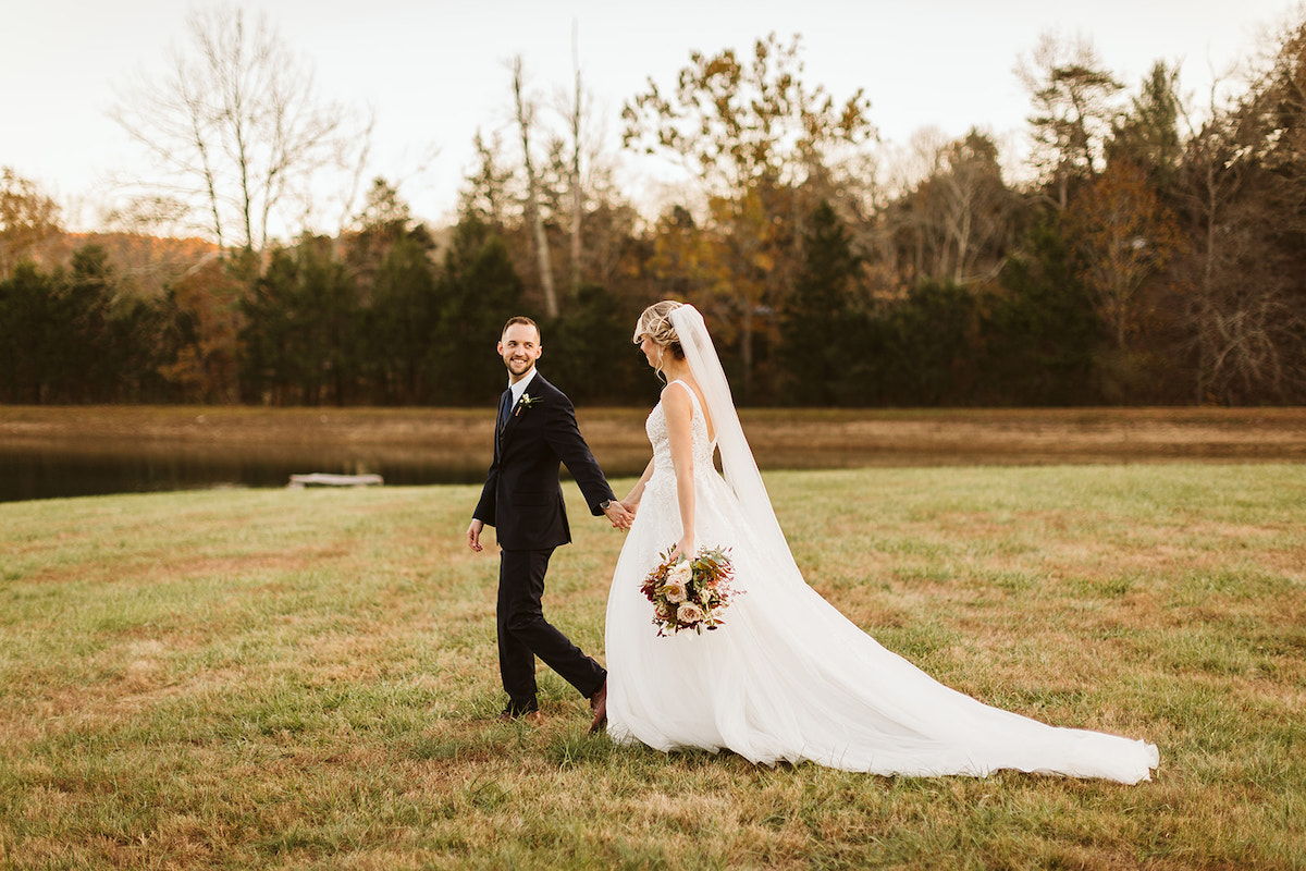 Bride and groom hold hands and walk through a grassy field at Cloudland Station's Lake Angela.