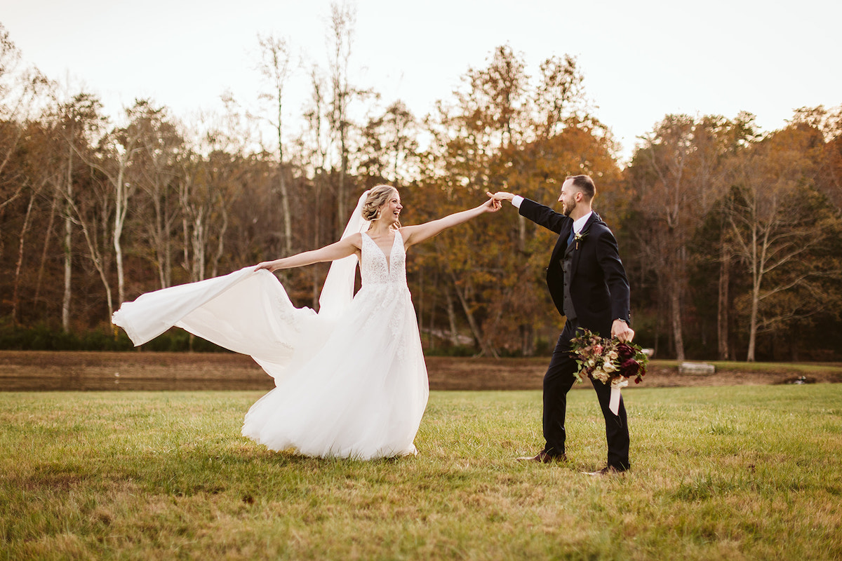 Groom twirls bride in a grassy field. He holds her bouquet by his side, and she holds her train as she spins under his hand.