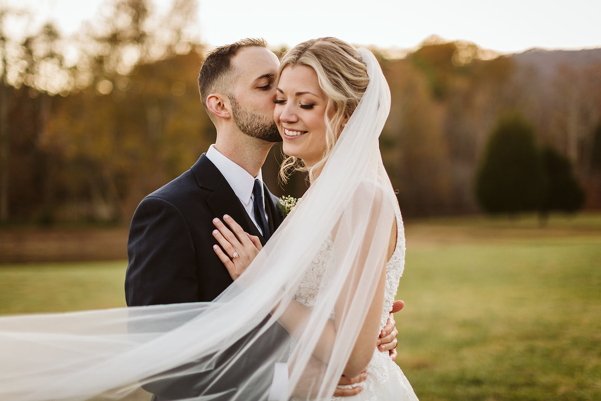 Groom holds bride around her waist and kisses her cheek as her veil streams around them