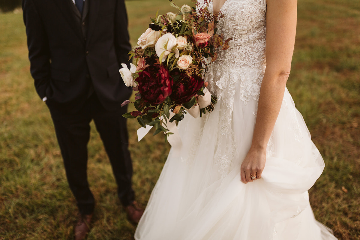 Bridal holds her Petaline bouquet filled with dark red flowers, pink and white roses, and greenery against her lace gown.