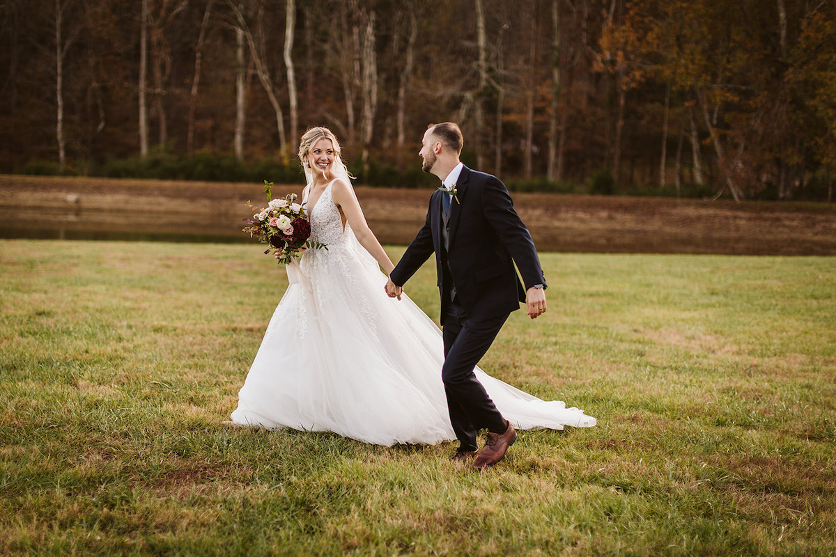 Bride and groom hold hands and walk through a grassy field at Cloudland Station's Lake Angela.