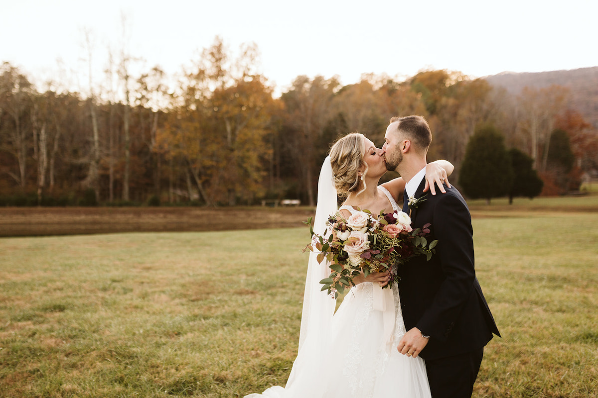 Bride and groom kiss in a grassy field. She has one arm around his shoulders and holds her large bouquet in the other