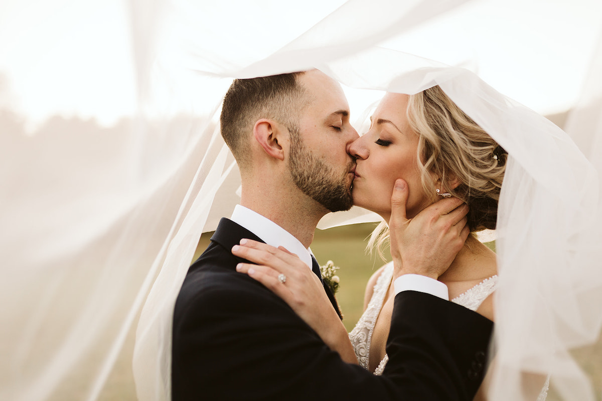 Bride and groom kiss under her veil, his hands cup her face and she holds his shoulder