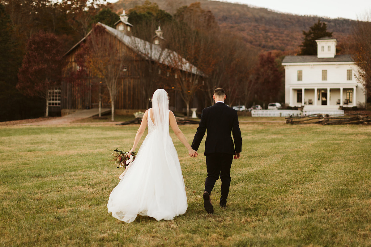 Bride and groom walk through grassy field toward Peacock Hall and the barn at The Homestead at Cloudland Station