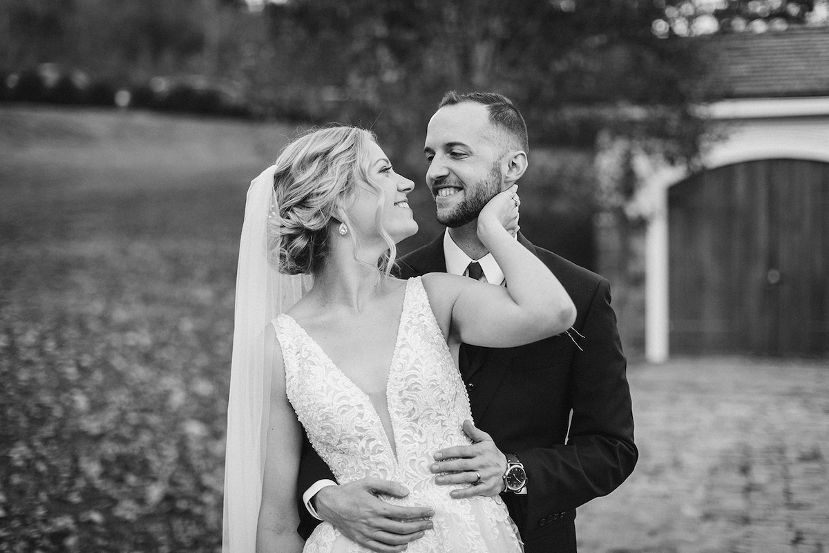 Groom stands behind bride with his hands around her waist. She looks back at him, her hand at his neck.