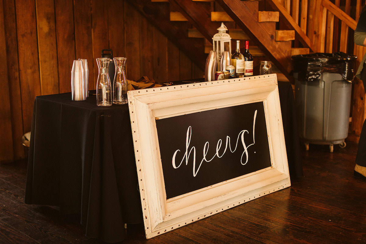 A large blackboard in a white frame with "Cheers!" written on it sits on the floor in front of a table with bottles of wine