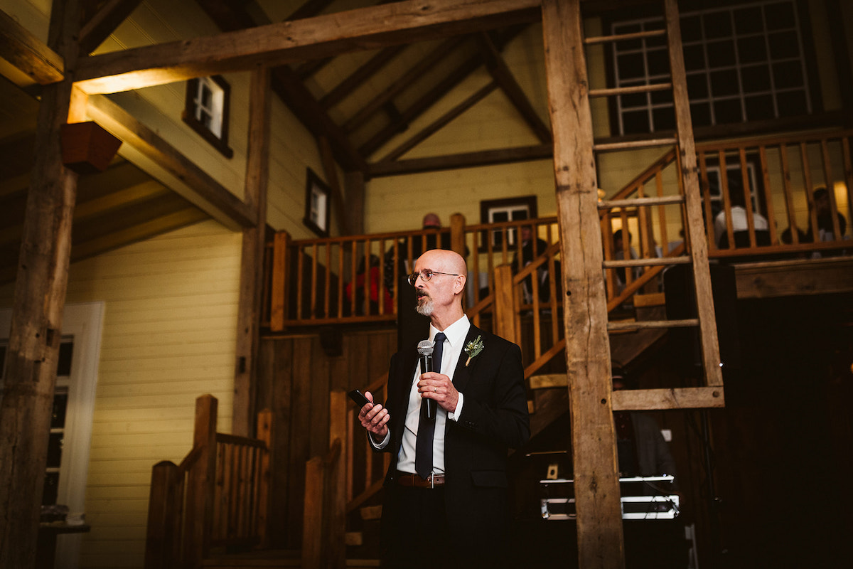 Man in dark suit and tie speaks into microphone under the exposed beam ceiling and loft of The Homestead at Cloudland Station