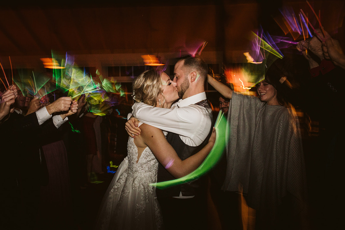 Bride and groom hug each other tightly and kiss while friends wave glow sticks around them