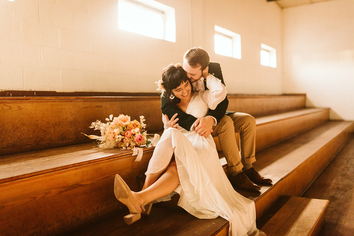 Bride and groom cuddle in the bleachers, her hand in his hair and her bouquet next to them.