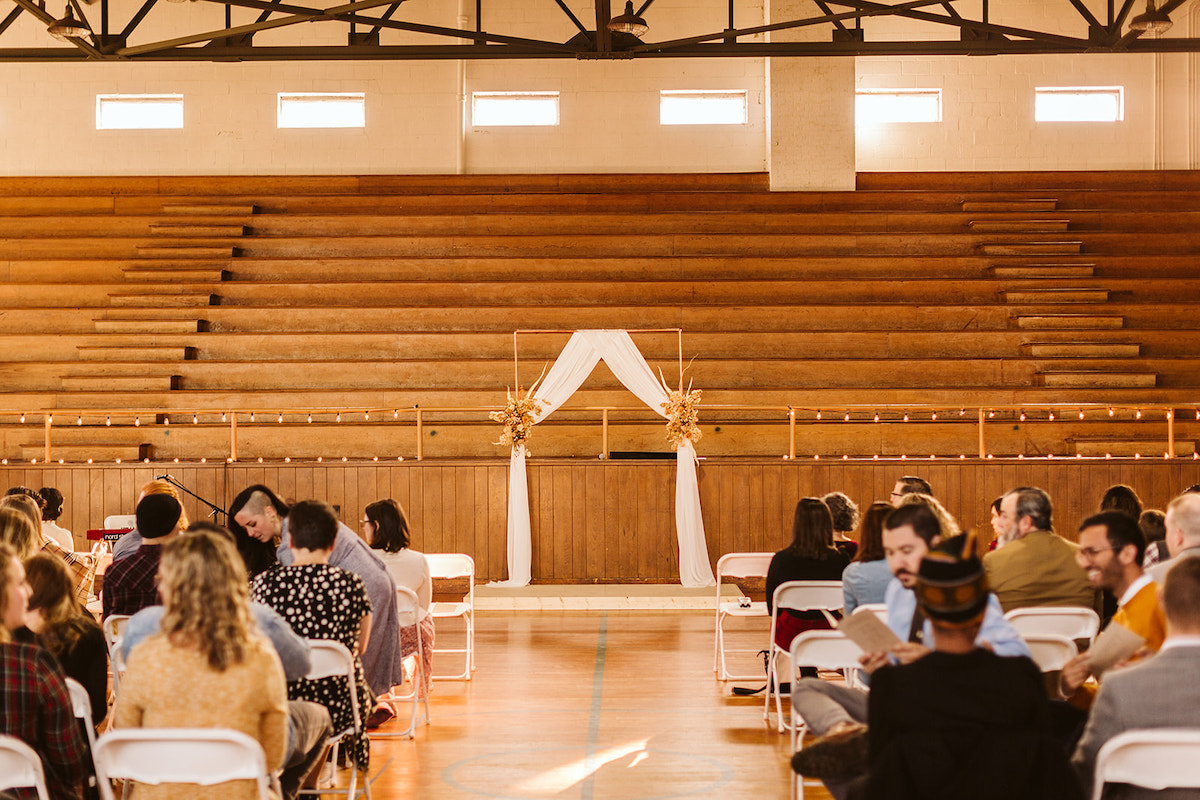 Simple copper pipe wedding arch draped with white linen in front of gymnasium bleachers