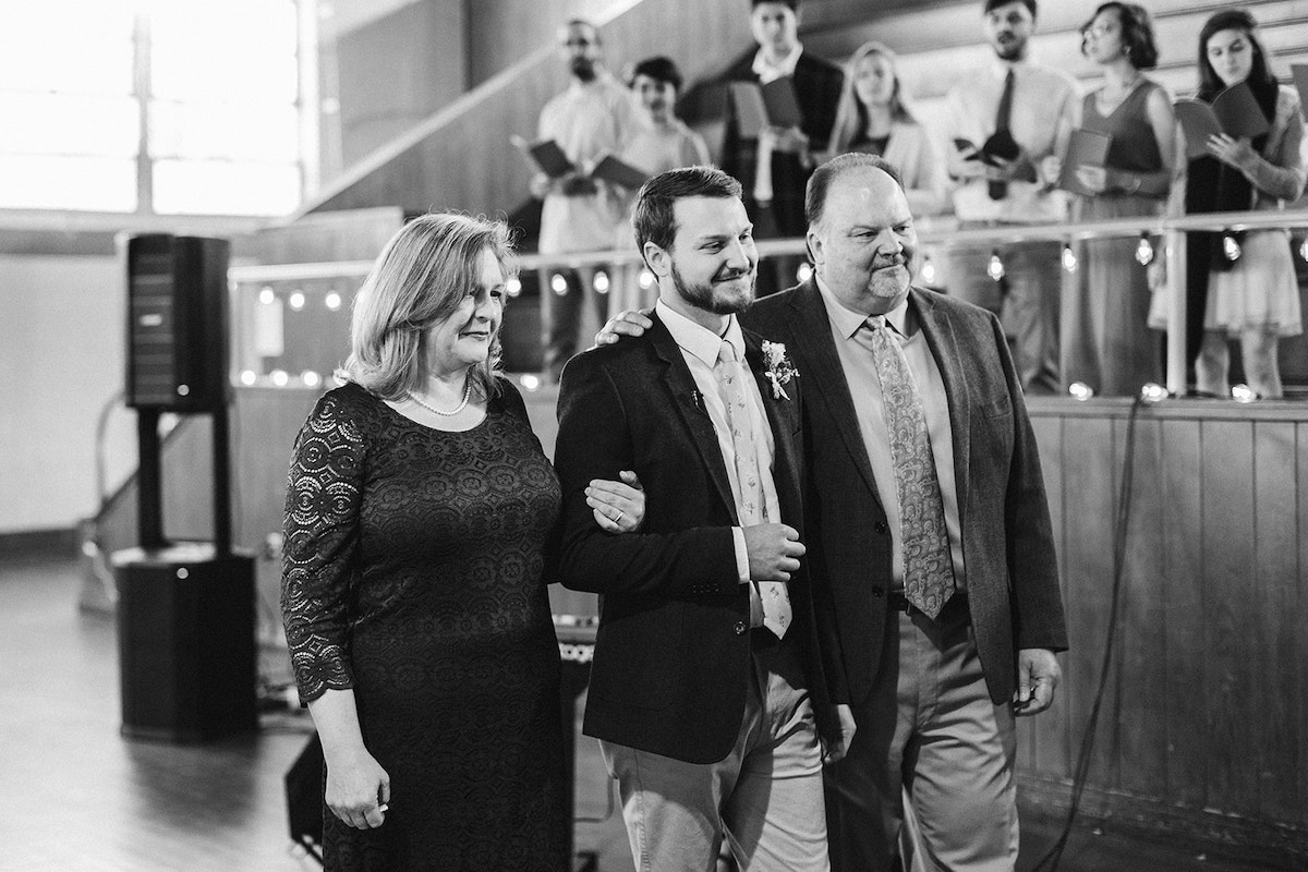 Groom walking arm-in-arm with his mother and father while friends sing in balcony behind him