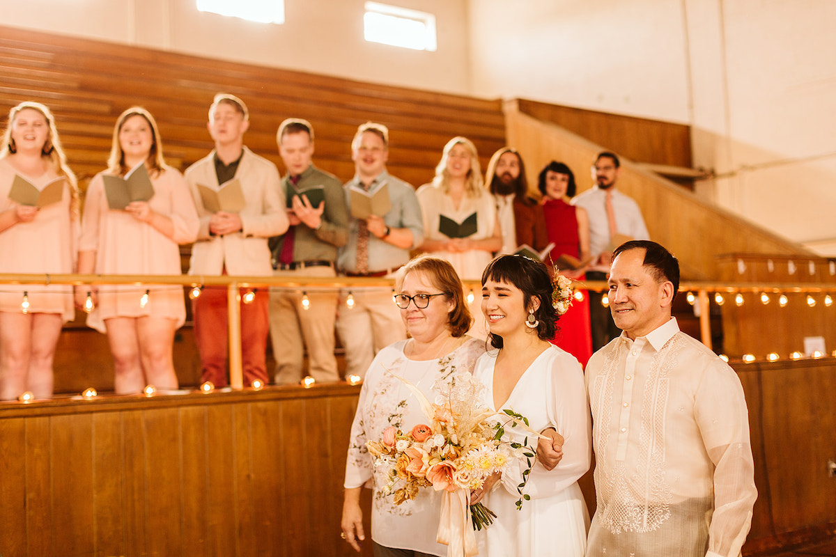 Bride walking arm-in-arm with her mother and father while friends sing in balcony behind her