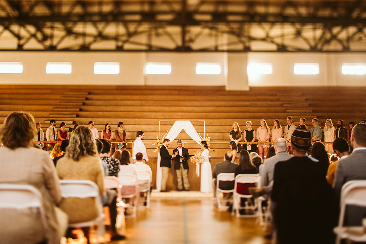 bride, groom, and minister stand under a simple copper pipe wedding arch in front of wooden bleachers in a gymnasium
