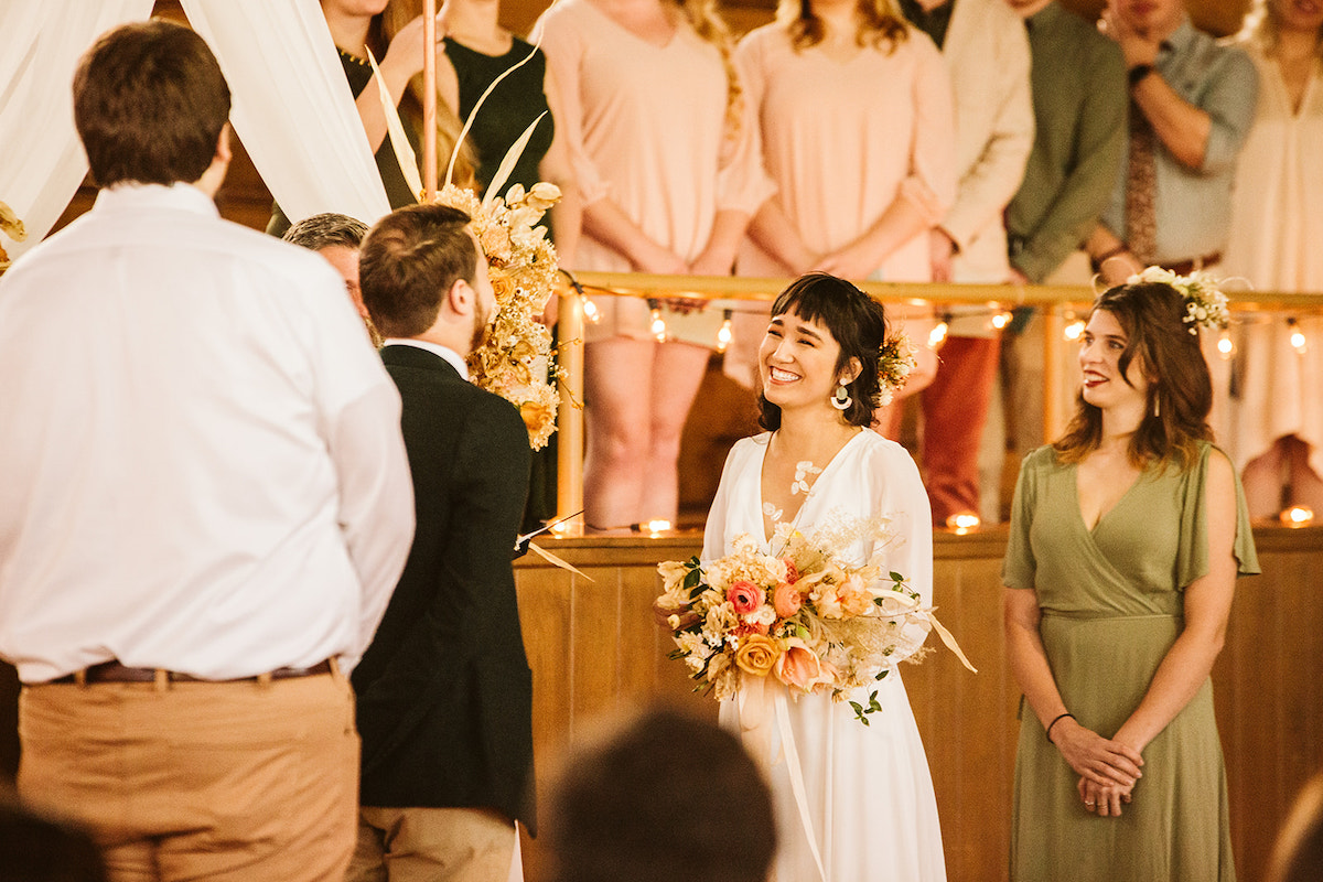 Bride smiles at groom, her maid of honor behind her and several wedding attendants watch from gym bleachers