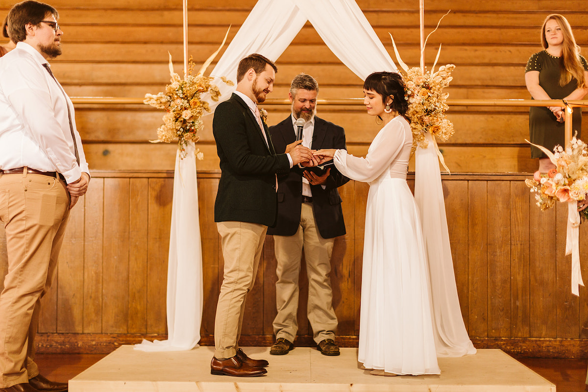 Groom slides a rind onto bride's finger while their minister reads from his book and wedding attendants watch