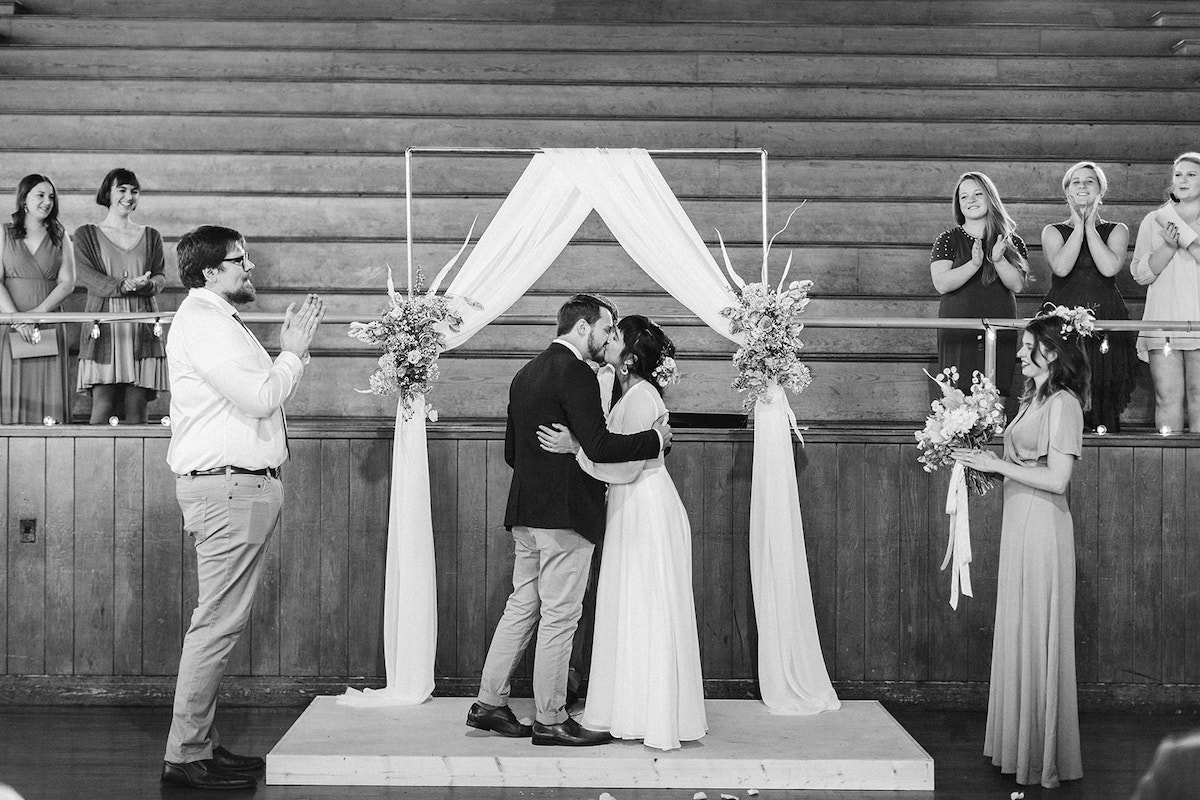 Bride and groom kiss beneath a simple copper wedding arch. Their wedding party looks on and claps.