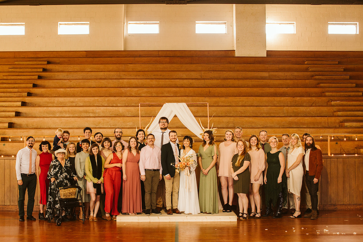 Bride and groom and their wedding part pose in front of wooden school gymnasium bleachers