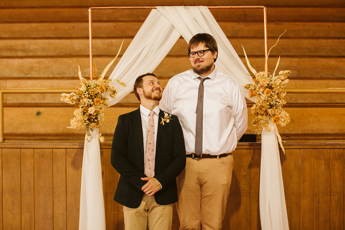Groom looks up at his much taller best man as they stand beneath a simple copper wedding arch