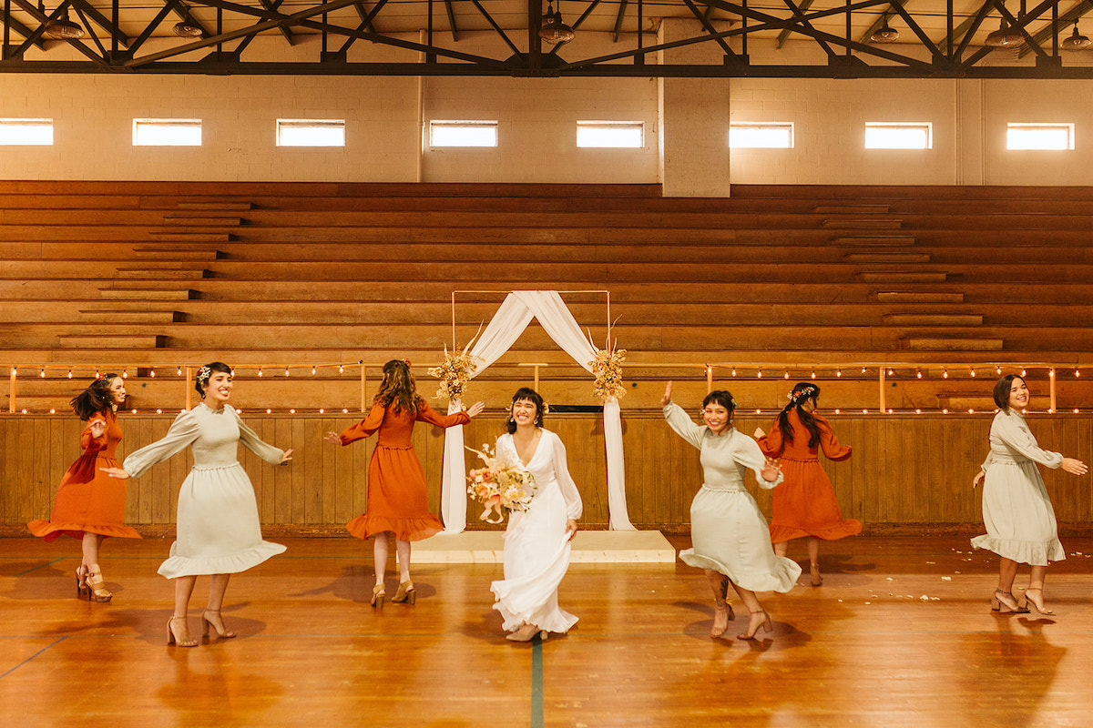 Bride and bridesmaids twirl in their dresses in front of simple copper wedding arch in front of wooden gymnasium bleachers