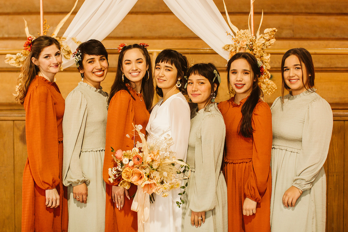 Bride and her bridesmaids in light green or amber-colored dresses stand beneath the simple, copper pipe wedding arch