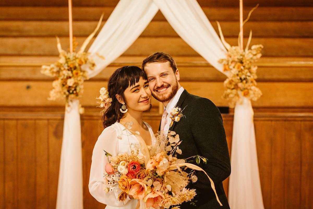 Bride and groom smile while holding her Southerly Flower Farm bouquet, their simple copper wedding arch in the background