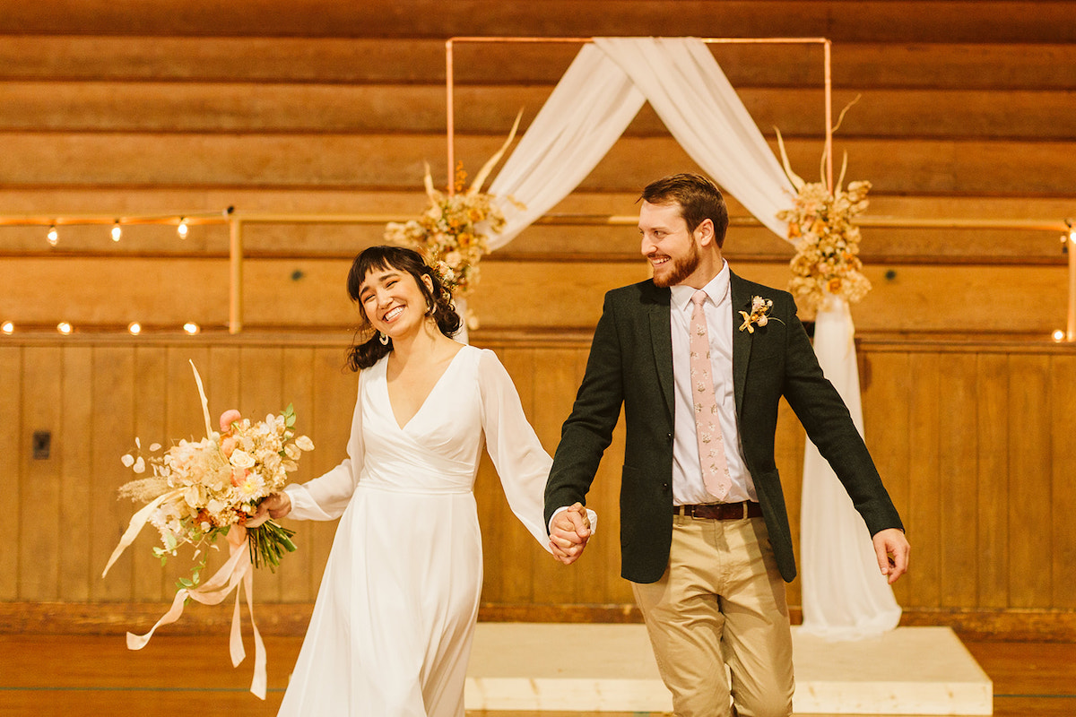 Bride and groom smile as they walk through a school gym, their simple copper wedding arch in the background