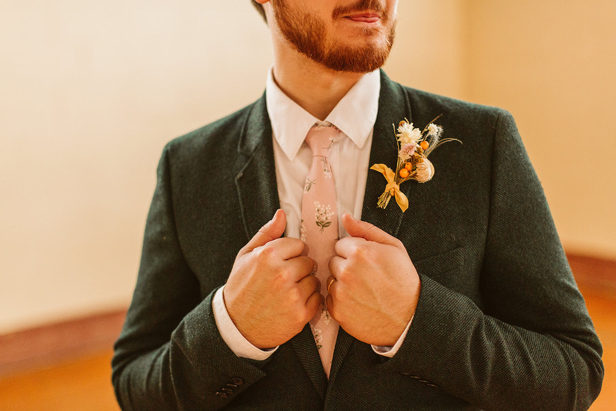 Groom holds lapels the of his suit coat. He wears a white shirt and pink tie with floral details.