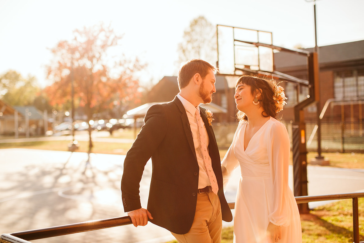 Bride in simple, long-sleeve wedding gazes at groom in the sunlight next to a basketball court