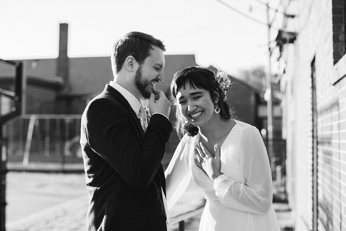 Bride and groom laugh together. She puts a hand on her chest, and he scratches his bearded chin.