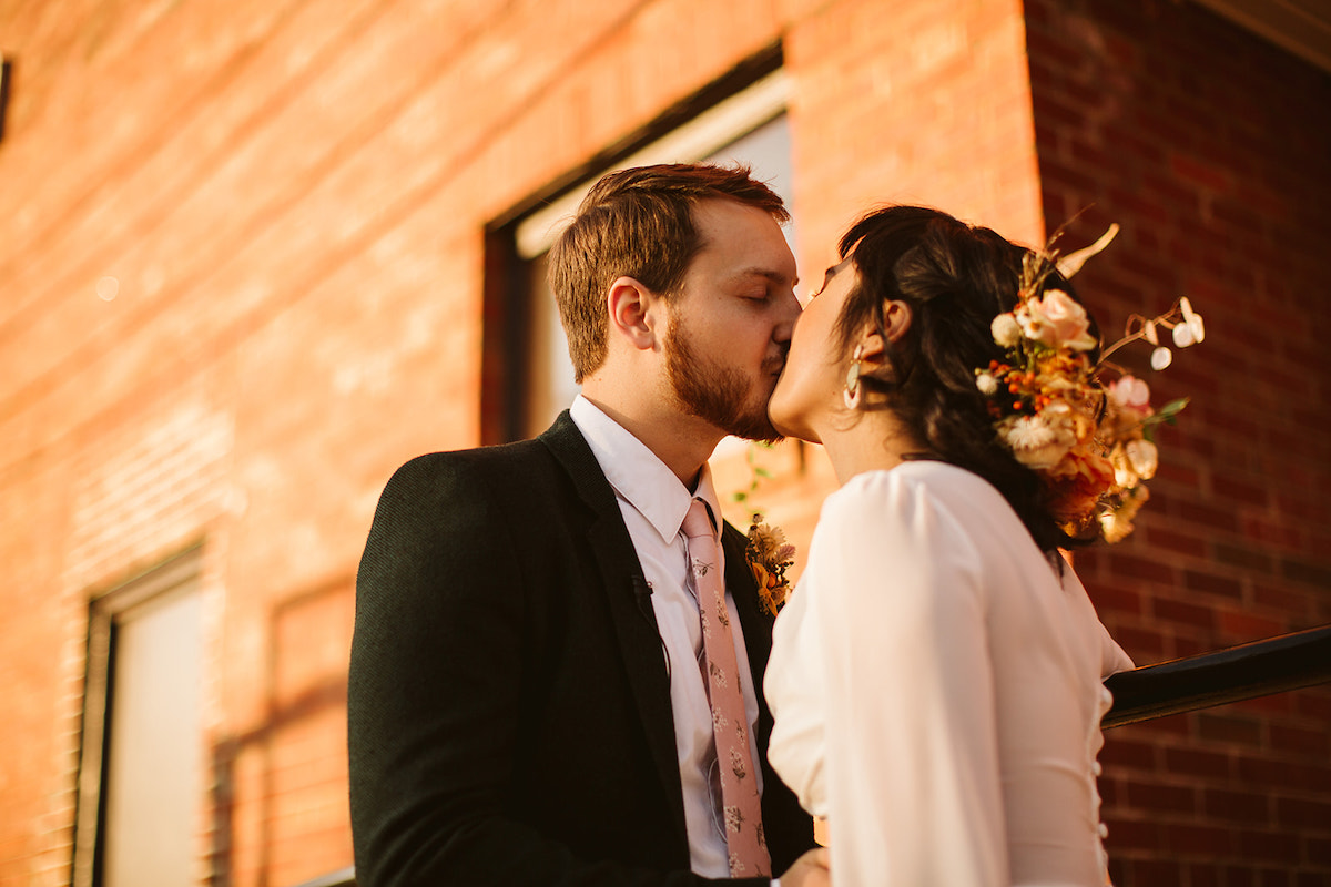 Bride and groom kiss next to a brick building. She wears a mixed Southerly Flower Farm bouquet in her hair.