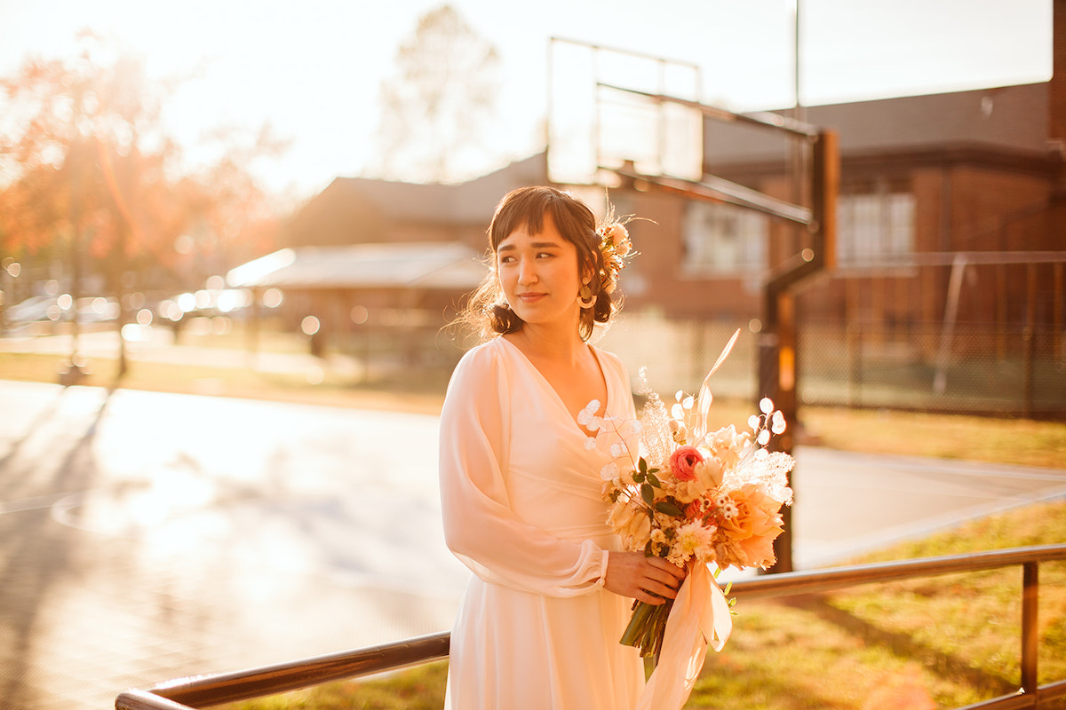 Bride in simple, long-sleeve wedding dress holds her bouquet at a basketball court