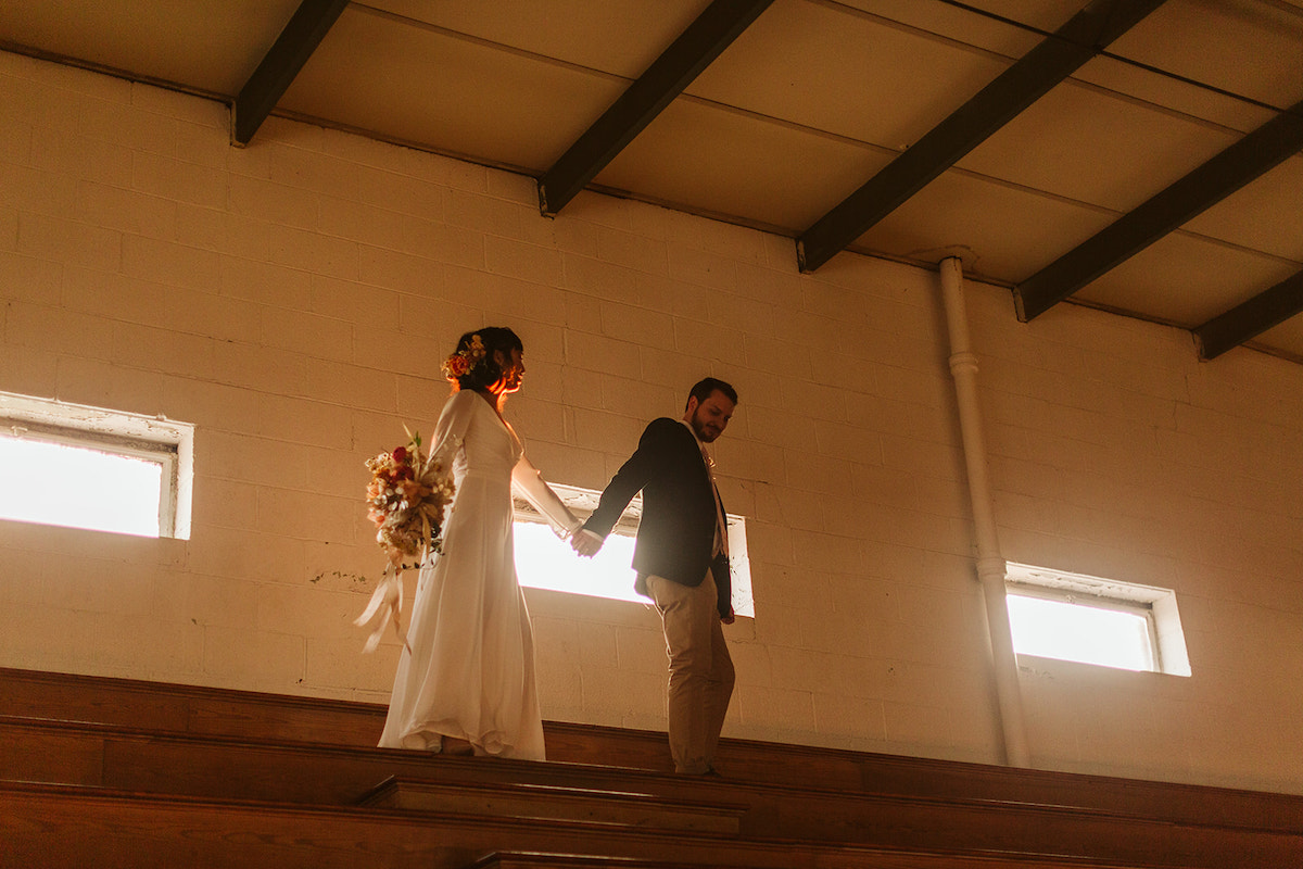 Bride and groom walk along the top row of wooden gymnasium bleachers
