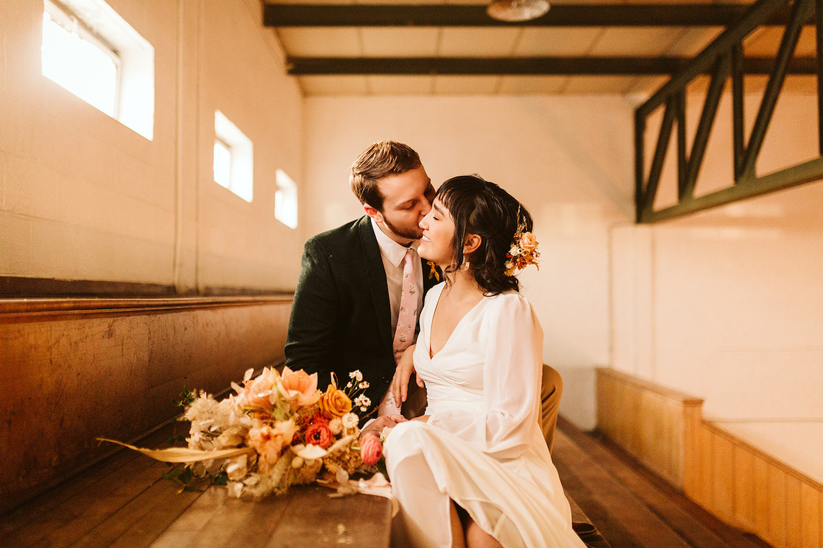 bride and groom sit kissing on wooden gymnasium bleachers with her Southerly Flower Farm bouquet next to them