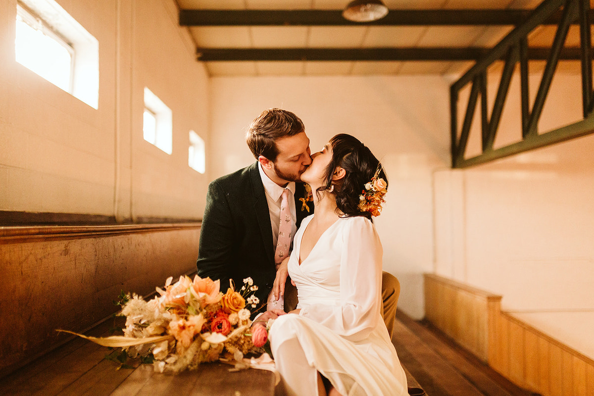 bride and groom sit kissing on wooden gymnasium bleachers with her Southerly Flower Farm bouquet next to them