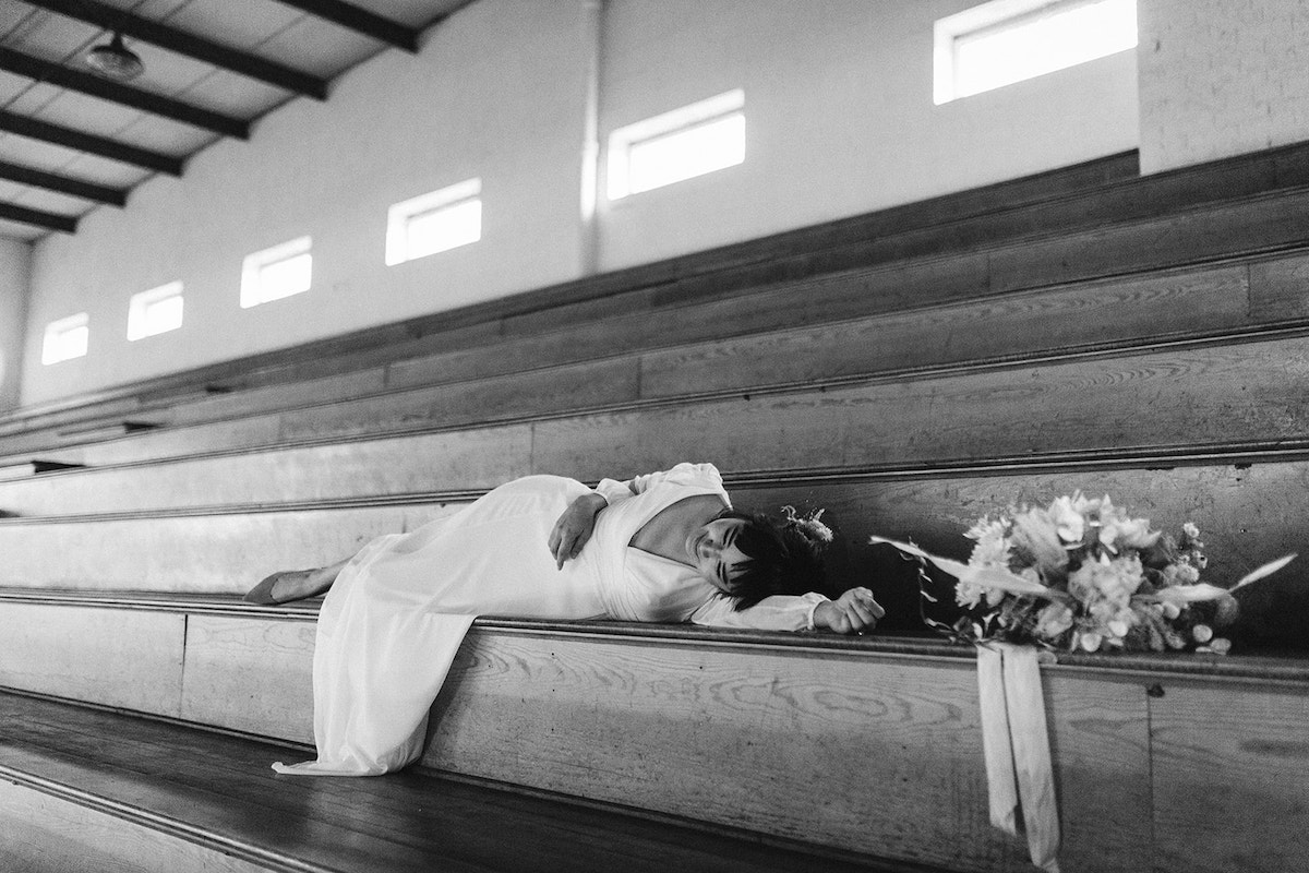 Bride in long-sleeve dress lies on wooden bleachers in school gymnasium, her bouquet next to her