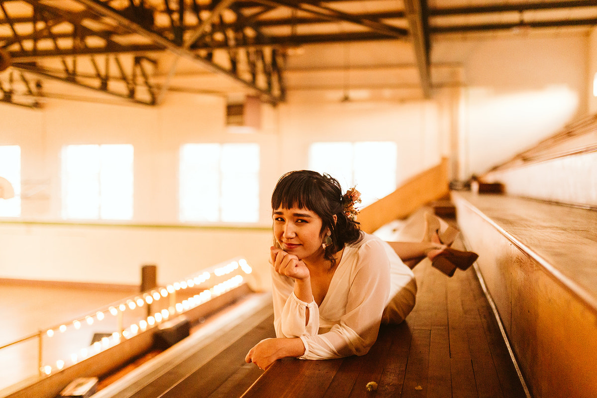 Bride in long-sleeve dress lies on wooden bleachers in school gymnasium