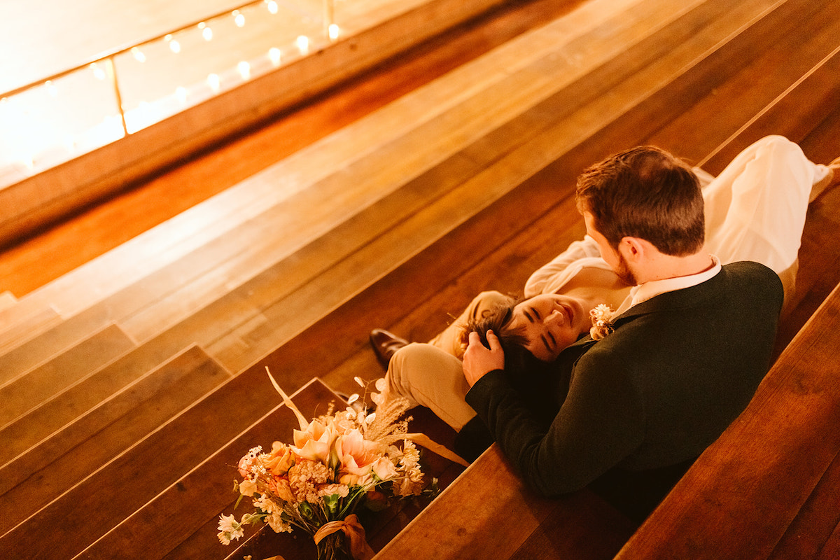 Bride lays with her head in groom's lap as he strokes her hair on wooden bleachers in a school gymnasium