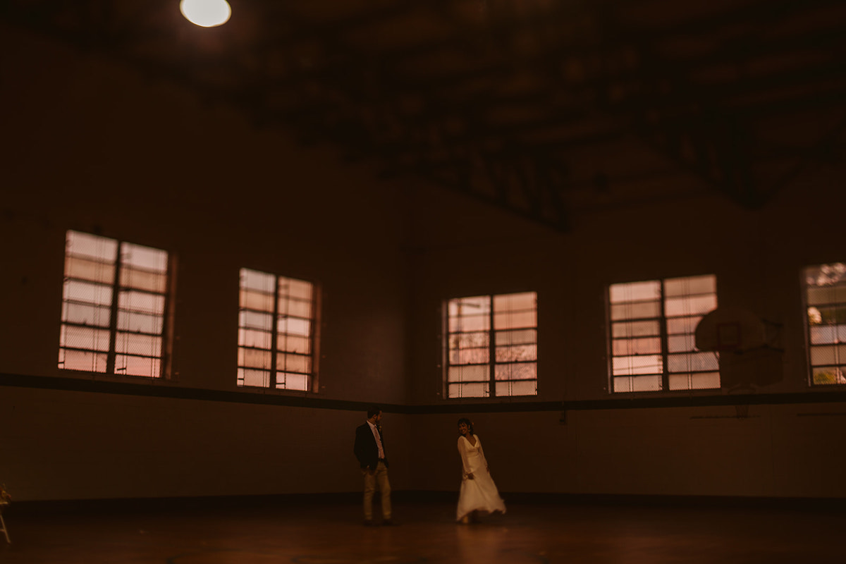 bride and groom look at each other under a basketball hoop in a shadowy gymnasium